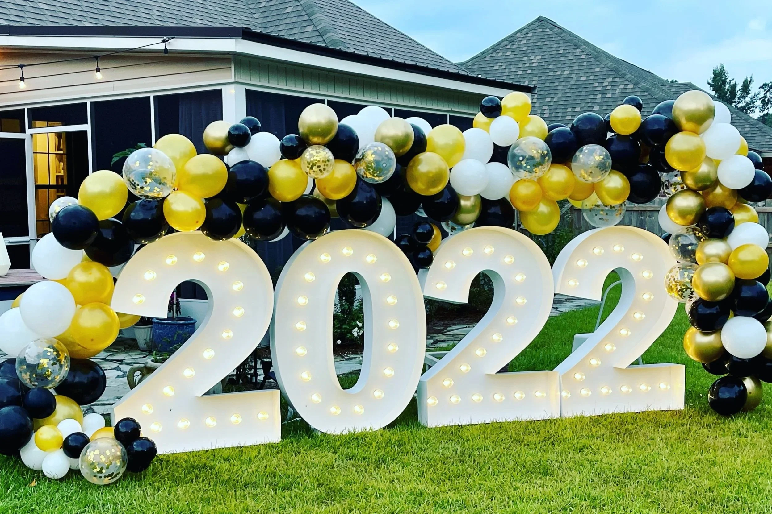 A decorative balloon display with black, gold, white, and clear balloons, including some with gold confetti, arranged around large white illuminated letters spelling out '2022,' outside a house with a green lawn, trees, and a covered porch in the background.