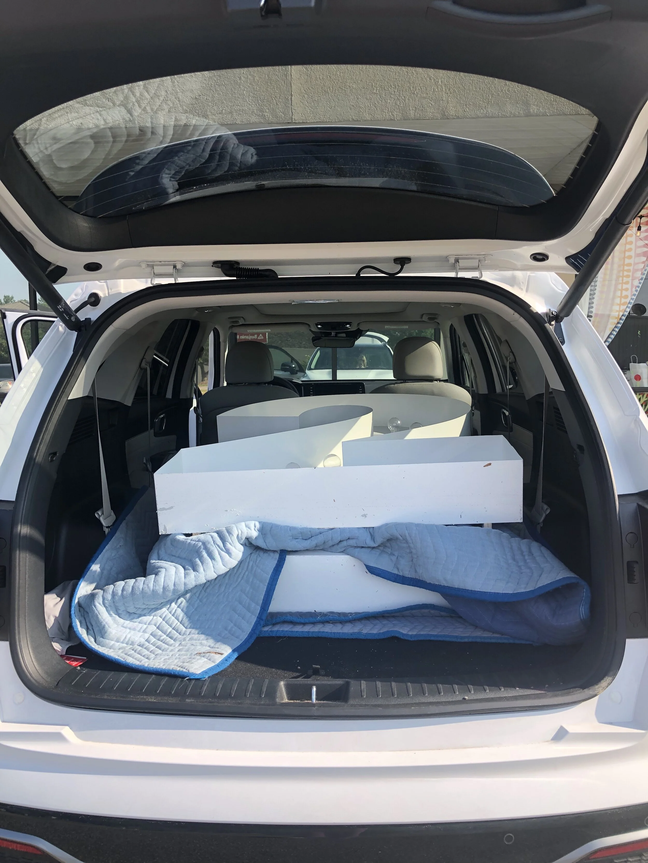 Rear view of an open car trunk with a white box and a blue quilted blanket inside, with some other items partially visible.