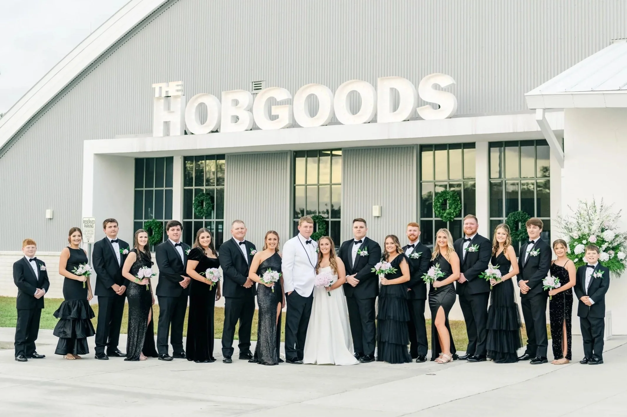 A group of wedding party members dressed in formal attire standing in front of a building with a sign that reads 'The HOBGOODS'.