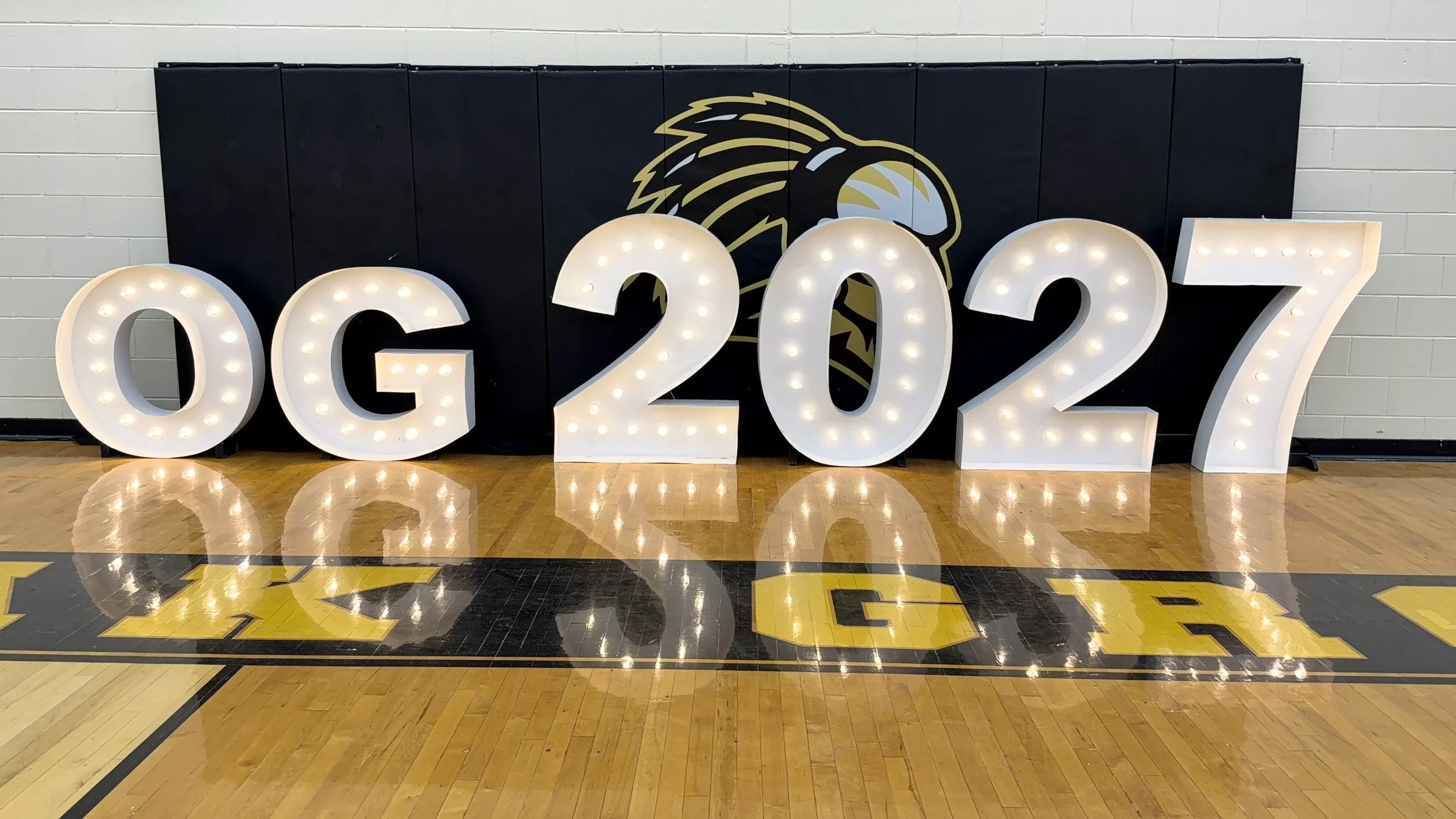 Illuminated large white letters spelling '2027' placed on a gymnasium floor with a black and yellow background featuring a stylized eagle head logo.