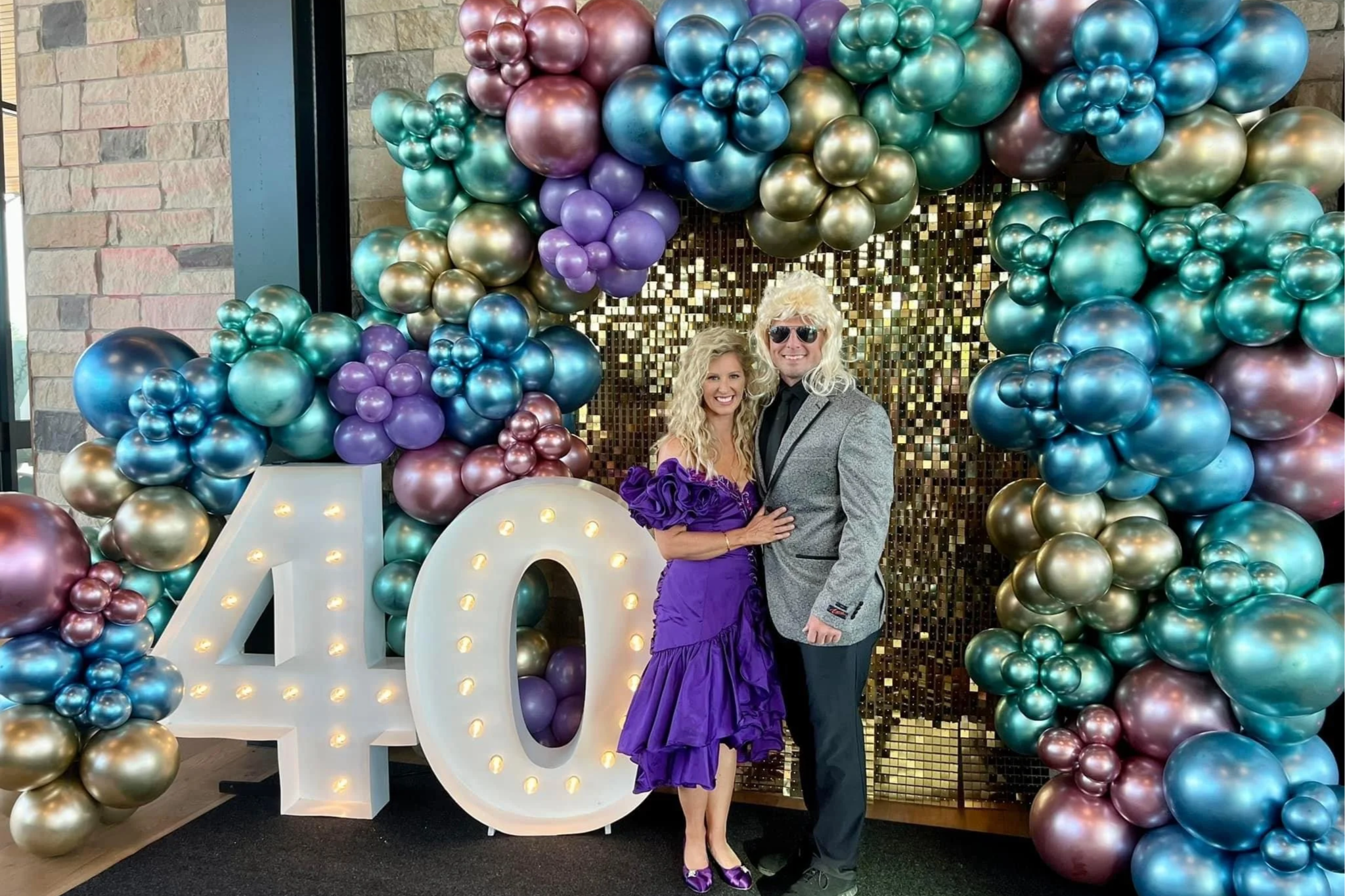 A couple dressed in formal attire standing in front of a decorative balloon backdrop with the number 40 lit up, celebrating a special occasion.