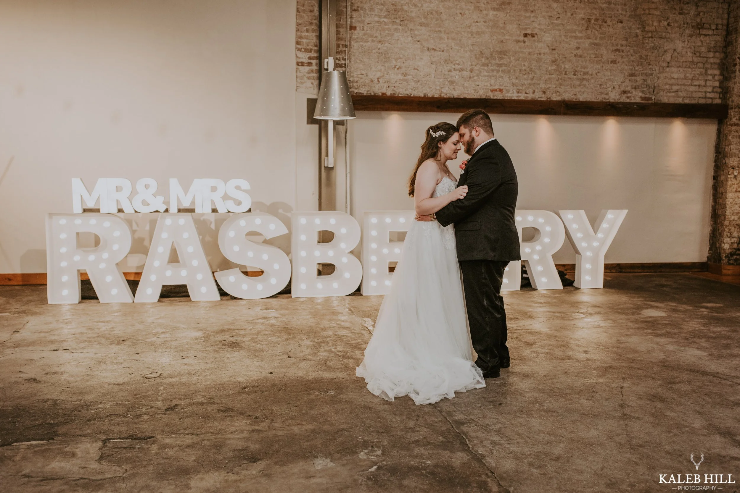 A bride and groom dance closely indoors in front of large illuminated letters spelling 'Mr & Mrs' and 'RASBERRY'. The background features exposed brick wall and modern lighting.