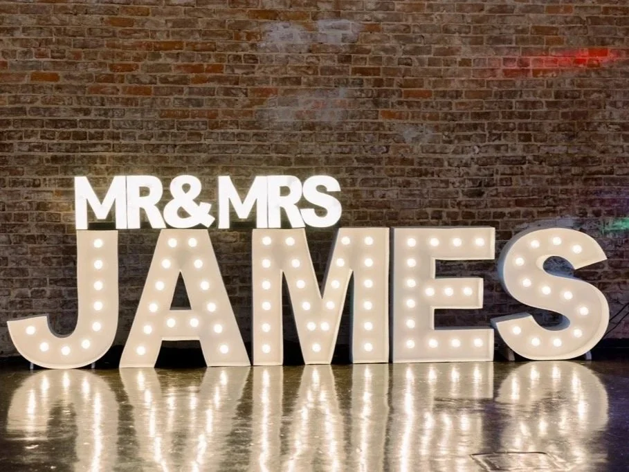 Large illuminated marquee letters spell out 'MR & MRS JAMES' against a brick wall at a wedding celebration.