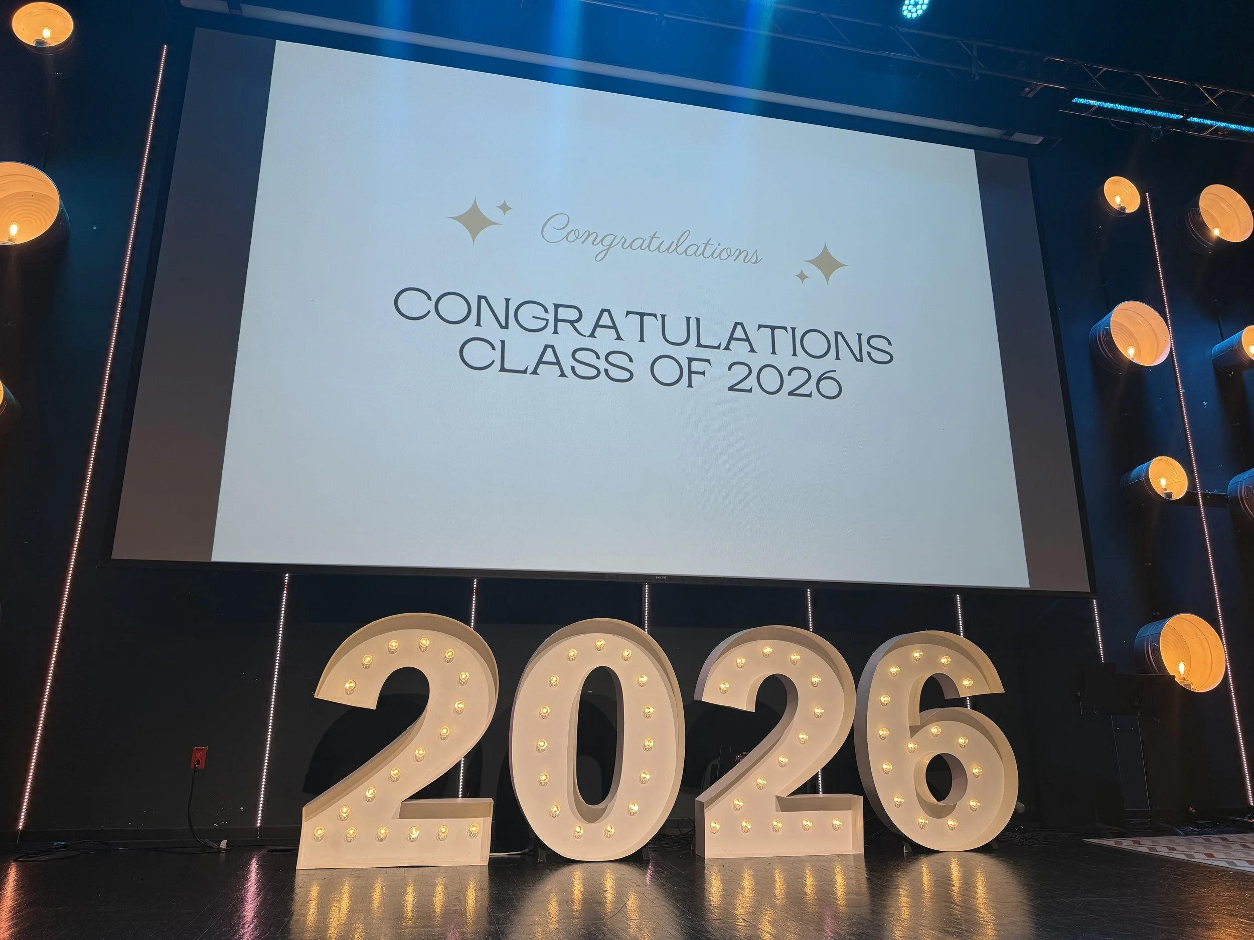 Large illuminated '2023' sign with graduation cap shape, decorated with small bulbs, set against a dark background, celebrating the graduating class of 2026.
