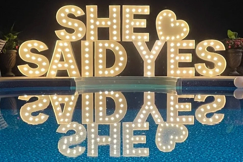 Illuminated marquee sign spelling "SHE SAID YES" with a heart, reflected in a pool of water, surrounded by potted plants.
