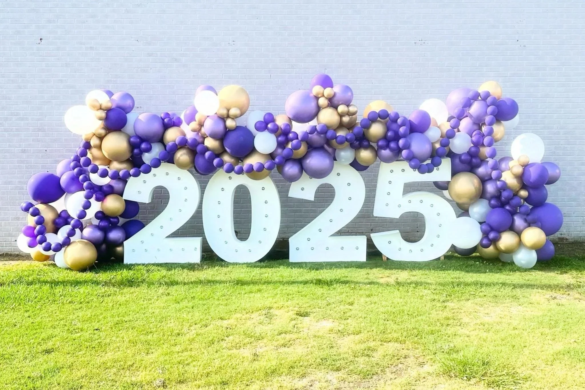 Decorative display of large white numbers '2025' surrounded by purple, gold, and white balloons arranged outdoors against a brick wall.