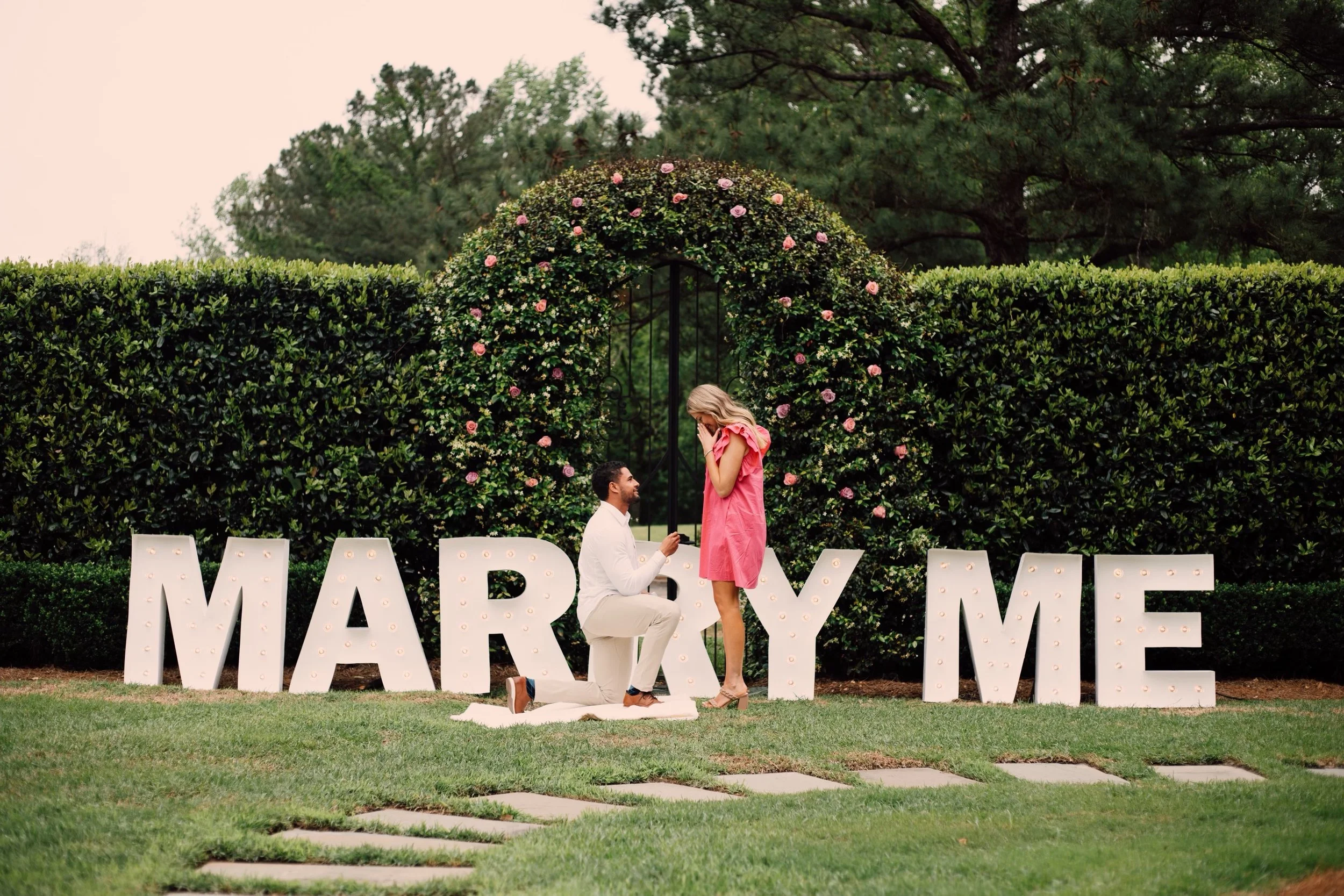 A couple participating in a marriage proposal outdoors near large illuminated letters spelling 'MARRY ME'. The man is on one knee proposing to the woman, who is standing with her hands over her face, in front of a green hedge and trees.
