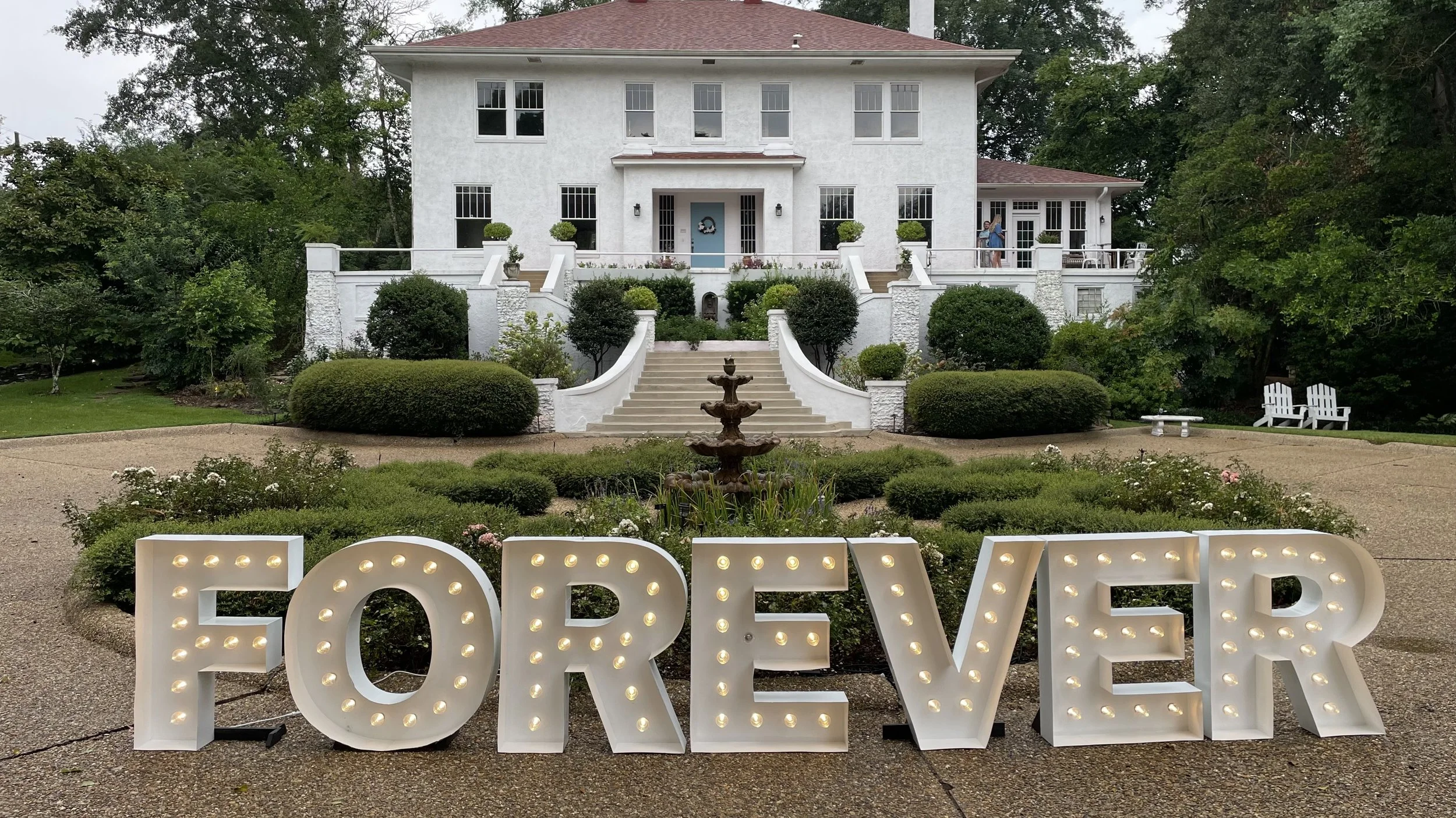 A large white house with multiple windows and a porch, surrounded by a well-maintained garden with bushes, trees, and a fountain. In the foreground, there is a large illuminated sign that spells out the word 'FOREVER' in decorative lighting.