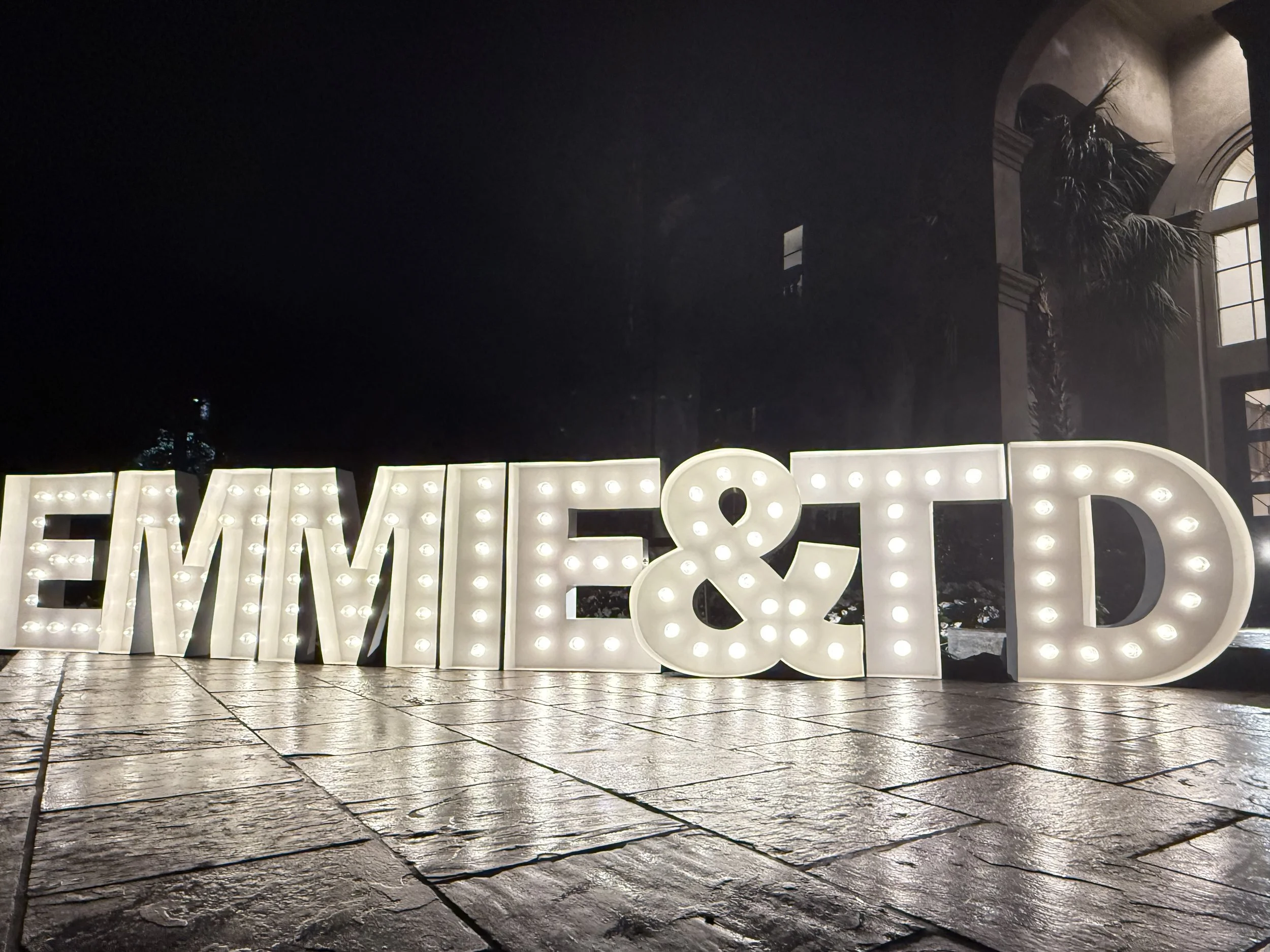 Large illuminated letter sign spelling out 'EMMIE & TD' on a tiled floor, with an arched window and a large plant visible in a dimly lit indoor space.