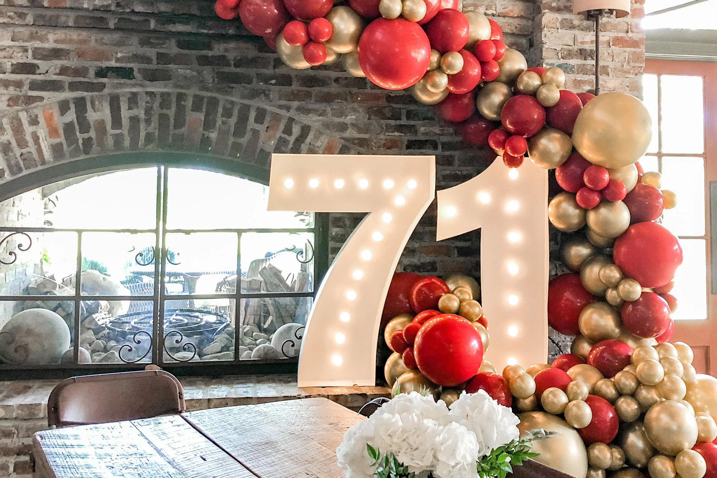 Decorative balloon arrangement with red and gold balloons, a large illuminated number '71', and a table with white flower centerpiece inside a brick-walled room with large window.