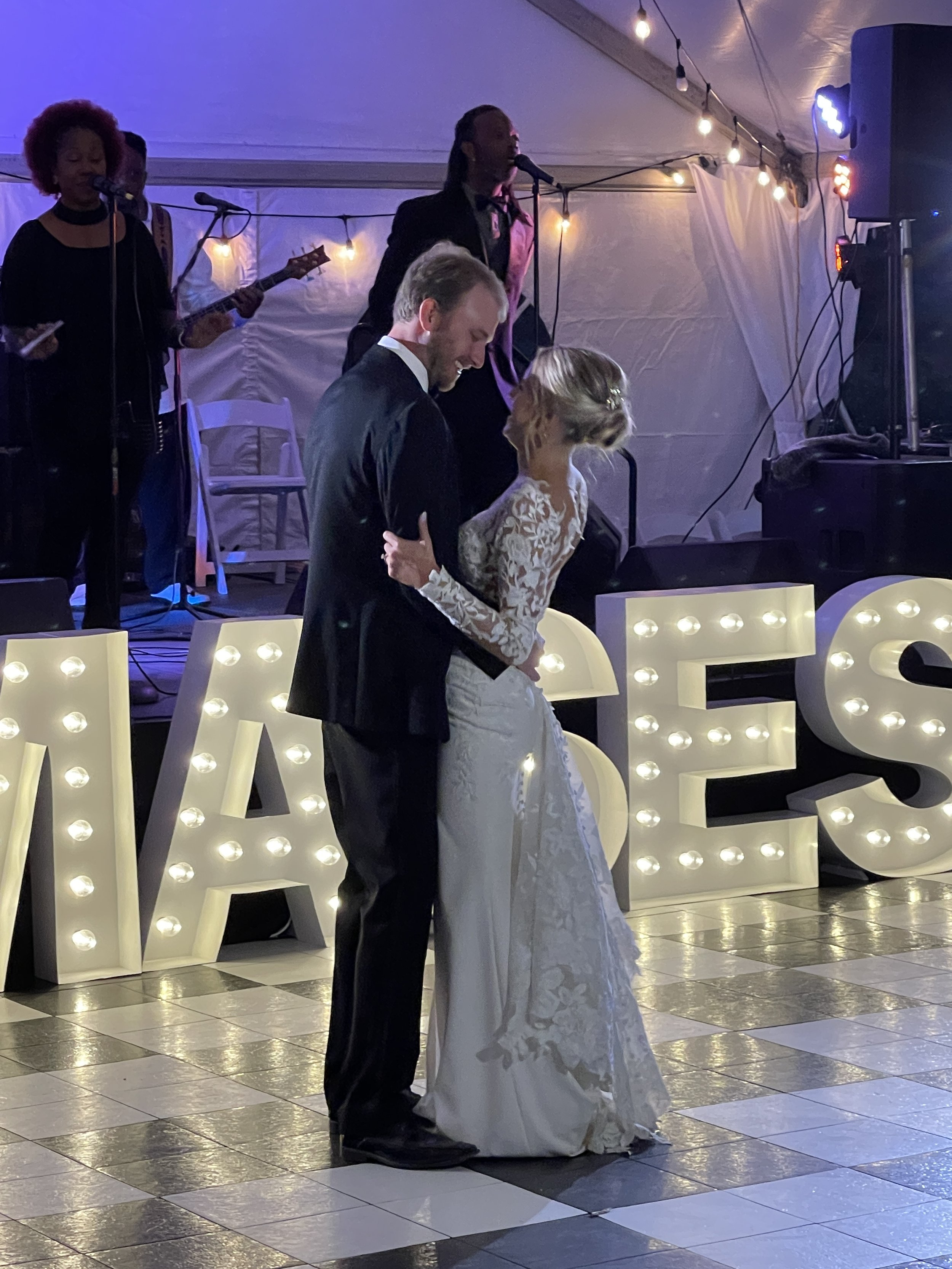 Bride and groom dancing at a wedding reception with live band in the background, illuminated letters displaying a message.