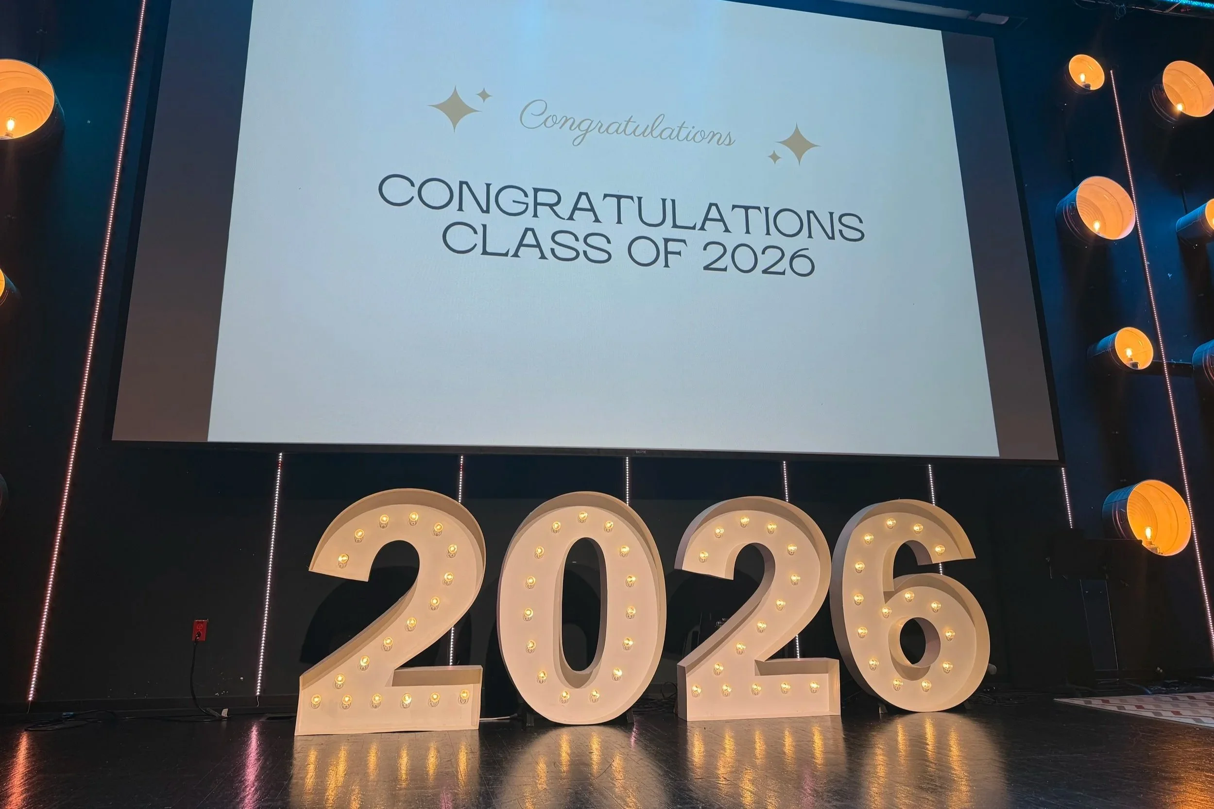 A large digital screen displaying the message "Congratulations Class of 2026" with decorative stars, surrounded by illuminated large marquee numbers '2026'.