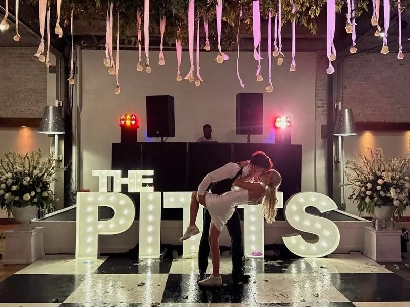 A couple is dancing and kissing in front of large illuminated letters spelling 'THE PITTS' at a wedding reception with floral arrangements, hanging ribbons, and DJ equipment in the background.