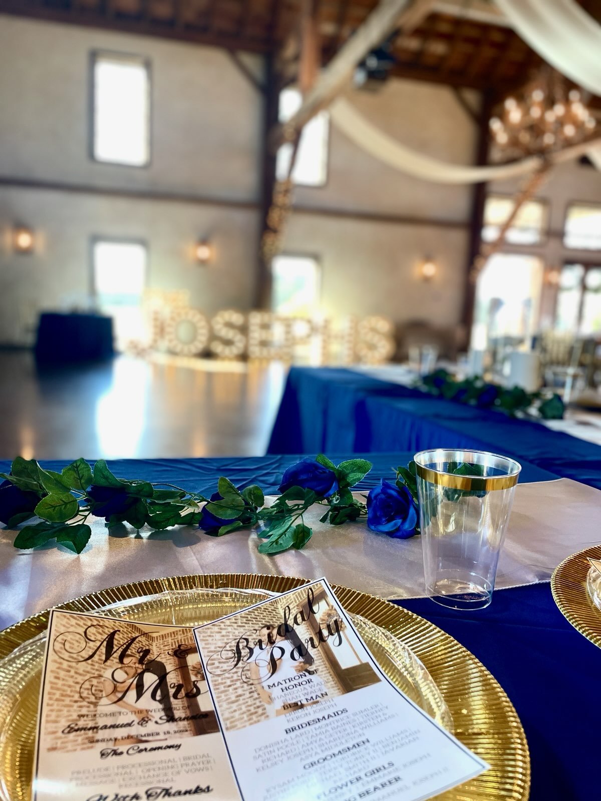 Wedding reception setup with a focus on a table setting featuring a gold-rimmed plate, a menu card titled "Mr. & Mrs." and "Bridal Party," a gold-trimmed plastic cup, and a blue and white floral arrangement. In the background, blurred letters spell "