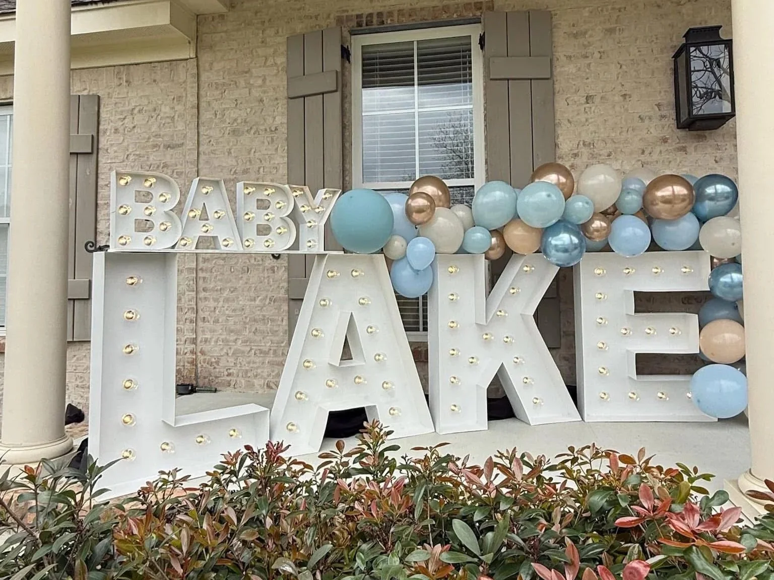 Decorative 'Baby LAKE' sign with white illuminated letters and pastel-colored balloons, set against a brick house with shuttered windows