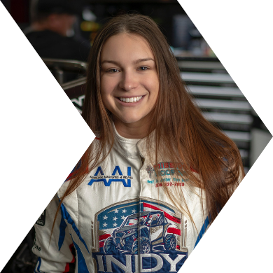 Young woman with long brown hair smiling in racing suit at race track.