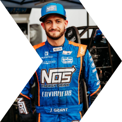 A male race car driver in blue racing suit and cap, standing in front of a race car.