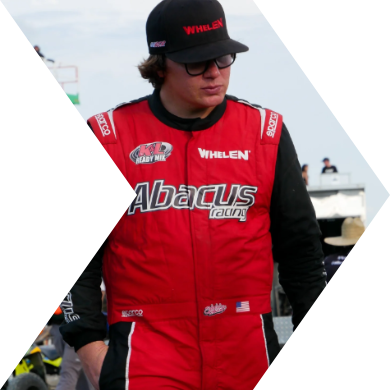 Person wearing a racing suit, cap, and sunglasses at a race track.