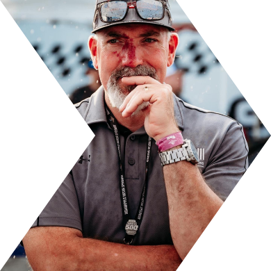 A man with a gray beard and mustache wearing a cap, sunglasses on his forehead, a gray polo shirt, and a watch, standing outdoors with a thoughtful expression.
