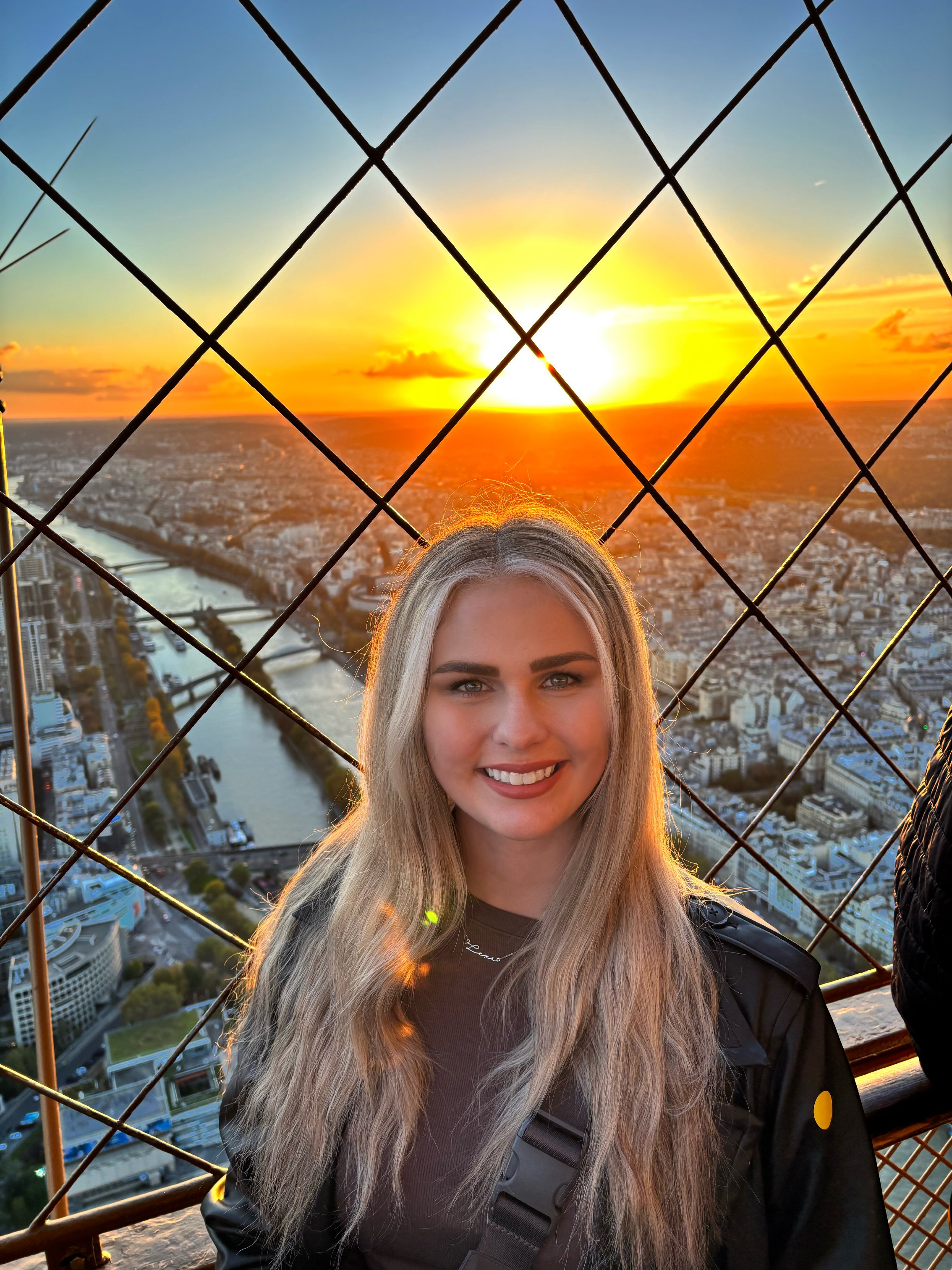 A young woman smiling in front of a cityscape during sunset on the Eiffel Tower observation deck, with a view of the Seine River and Paris buildings.