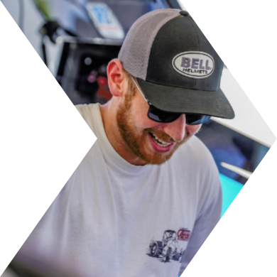 A man wearing a gray and black Bell helmet hat, sunglasses, and a white T-shirt smiling and looking down.