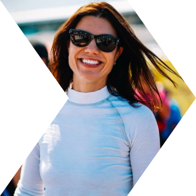 Smiling woman with long brown hair wearing sunglasses and a white long-sleeve top outdoors.