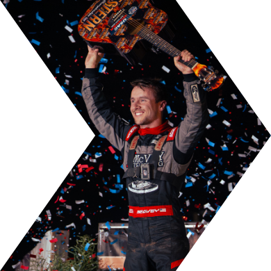 A race car driver is celebrating by holding a trophy above his head while standing on the podium with confetti falling around him.
