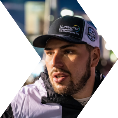 A young man with a beard wearing a black and white Nurtecon cap and a jacket, speaking or listening with a blurred background.