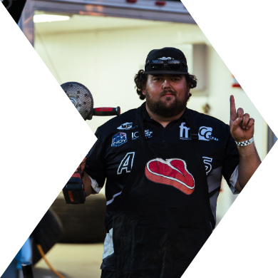 Person with a beard and curly hair wearing a black racing shirt and cap, holding a hockey stick, showing a peace sign with his right hand.