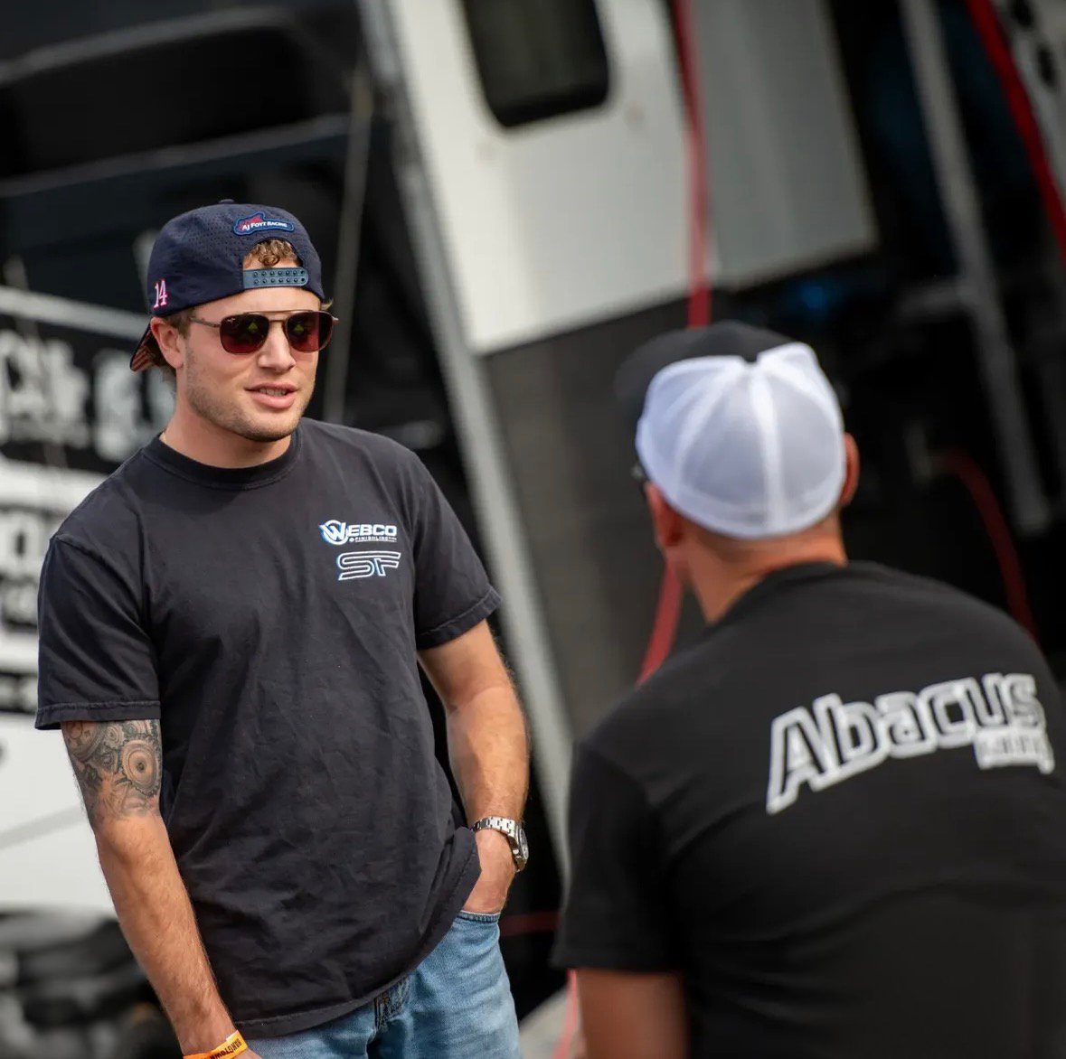 Two men having a discussion outdoors near race cars, one wearing sunglasses, a cap backwards, and a black t-shirt, and the other with a white cap and black shirt with white lettering.