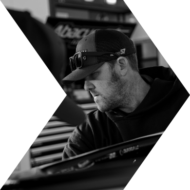 A man wearing a baseball cap, sunglasses on the brim, and a hoodie, working on a motorcycle in a workshop.