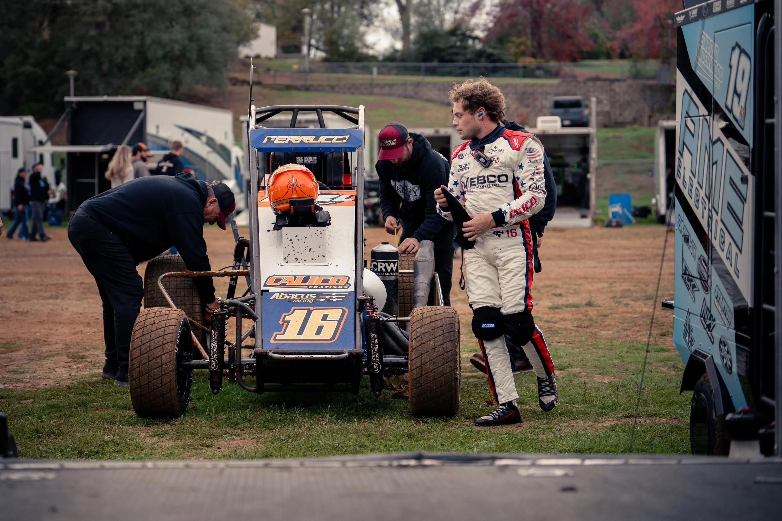 A race car driver in a white racing suit standing next to a small open-wheel race car with the number 16 on it, surrounded by mechanics and team members in a paddock area at a race track, with trailers and spectators in the background.