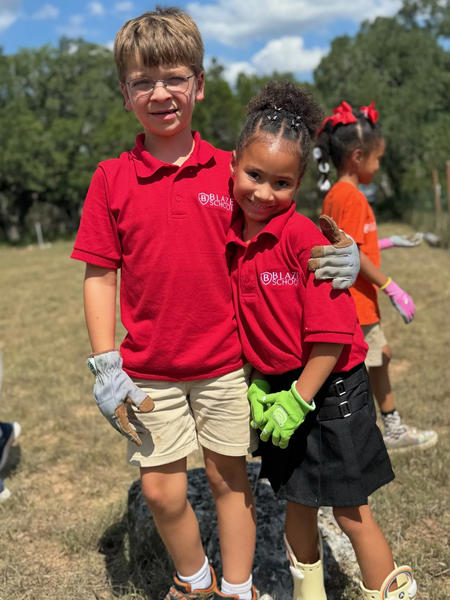 Two children wearing red Blaze School shirts, standing outdoors on a grassy field, smiling and hugging each other, with gloves on their hands.