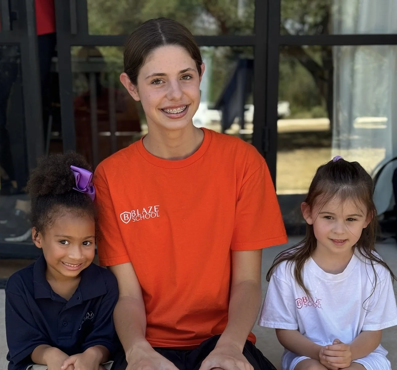 A young woman with braces and short brown hair, wearing an orange Blaze School t-shirt, sitting outside with two young girls, one with curly hair and purple bow, the other with straight brown hair, both wearing Blaze School t-shirts, smiling in front of a glass building.