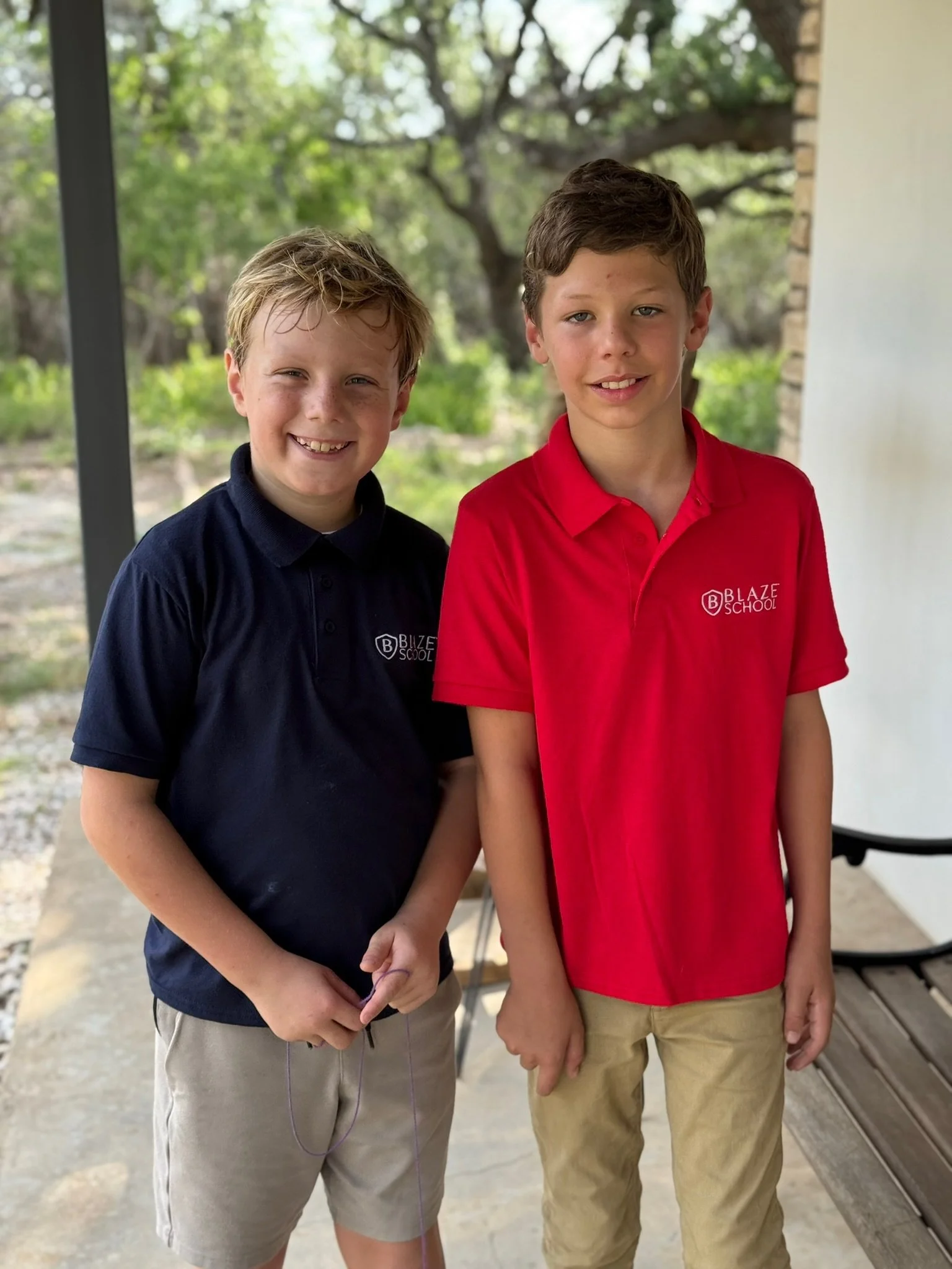 Two boys standing outdoors, smiling at the camera, dressed in school uniforms, with trees and a porch in the background.