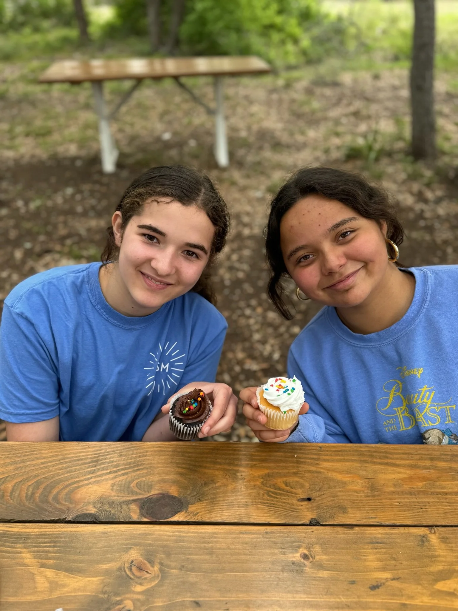 Two smiling girls sitting at a wooden picnic table holding cupcakes, outdoors in a wooded area.