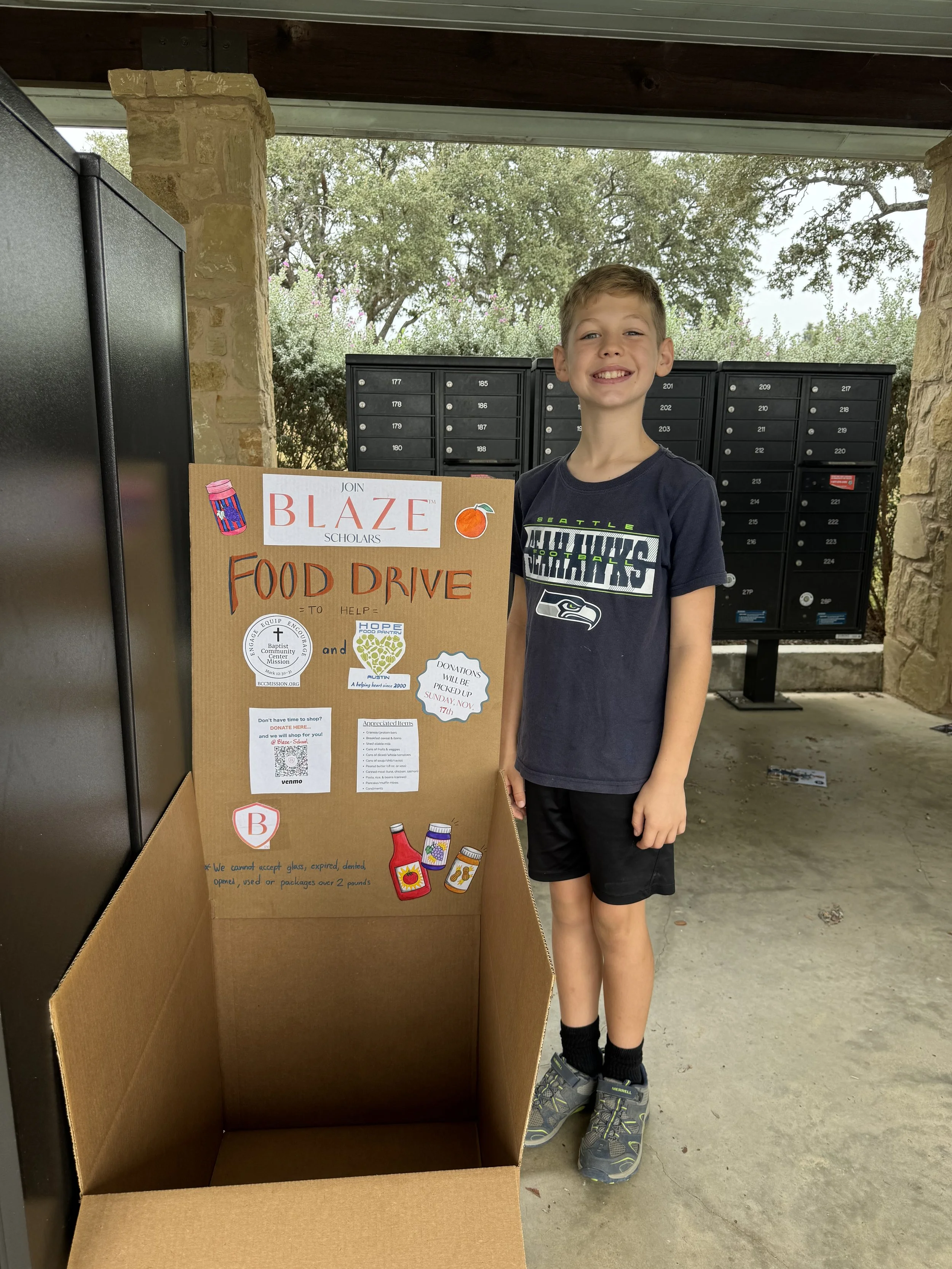 A young boy smiling next to a food donation box for a food drive organized by Blaze Scholars, with mailboxes and trees in the background.