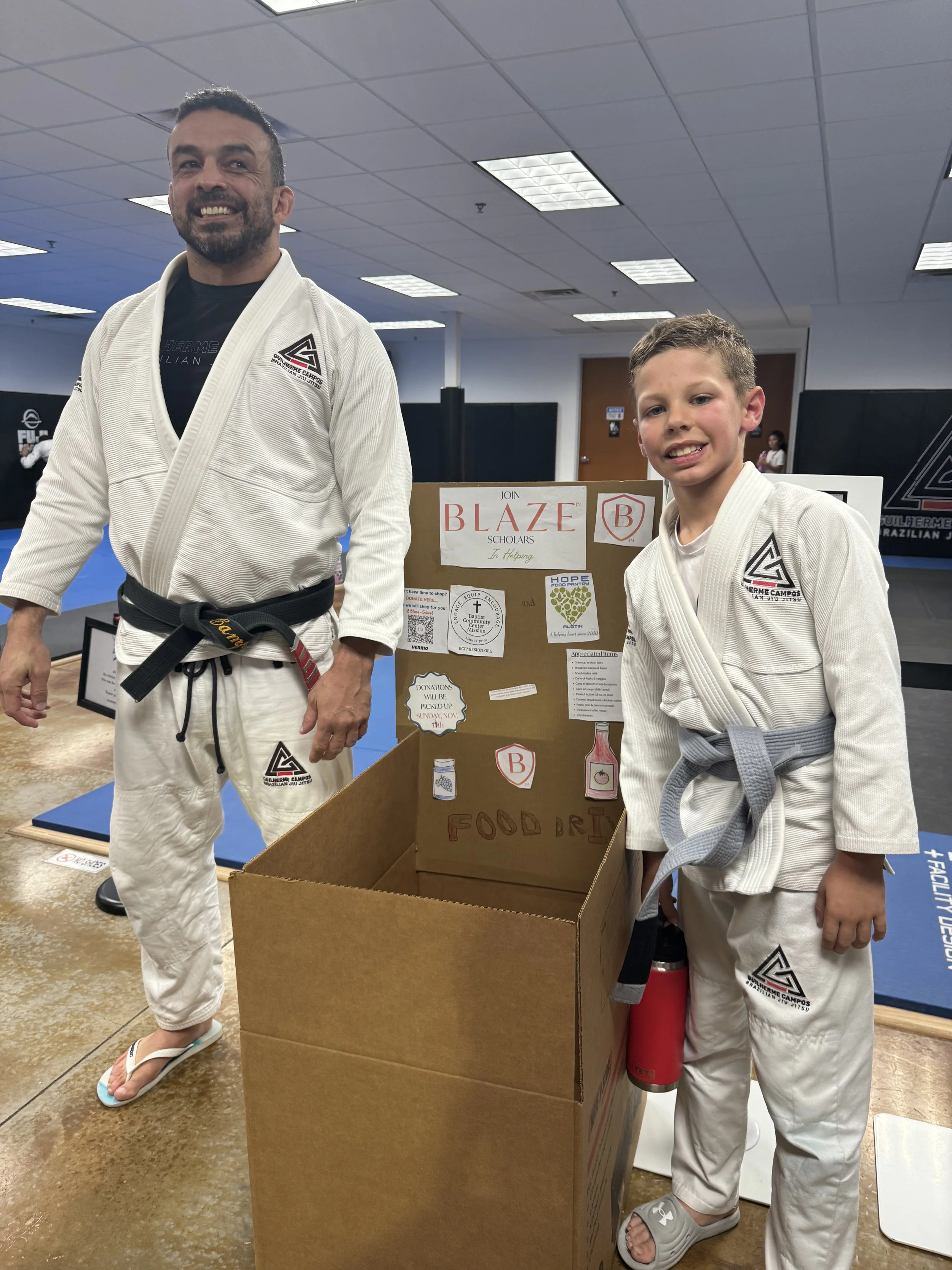 Two males in martial arts uniforms standing next to a donation box at a martial arts event. The older man has dark hair and a beard, wearing flip-flops, while the younger boy has short hair, wearing sandals. There is a sign on the donation box that reads 'Join Blaze Scholars' with illustrations and text related to food donations.