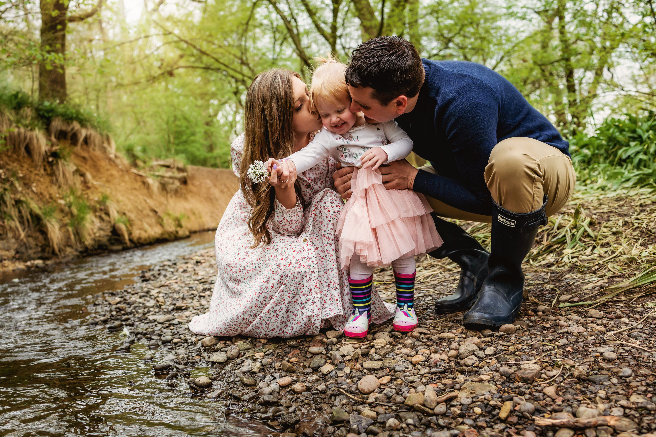 Family sat by a stream parents kiss little girl on the cheek while she smiles and wriggles during a family portrait session in Derby