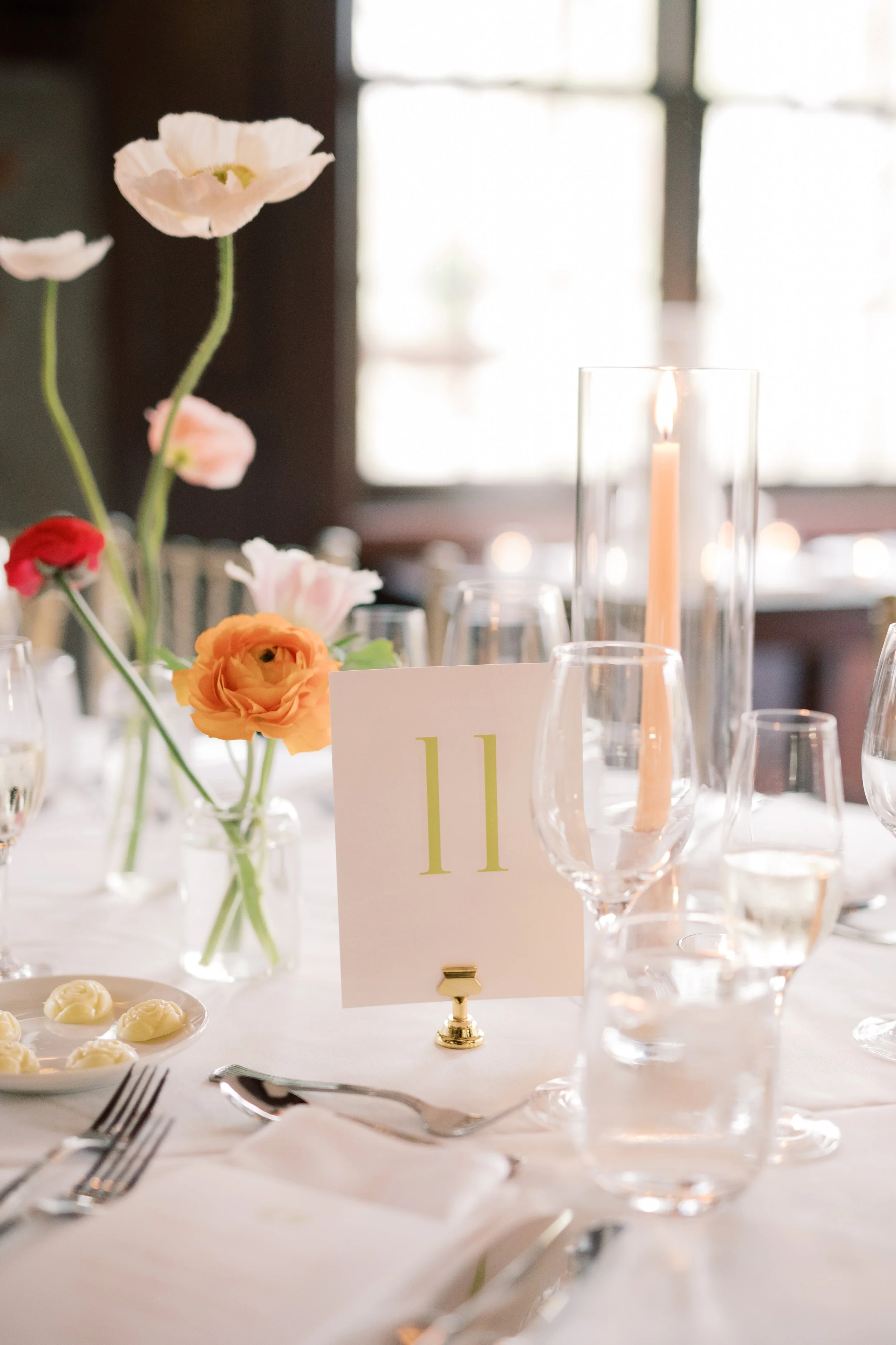 A table set for a formal event with flowers, a candle in a tall glass holder, glasses of water, a plate of butter, and a white tablecloth. A card with the number 11 is displayed on the table.