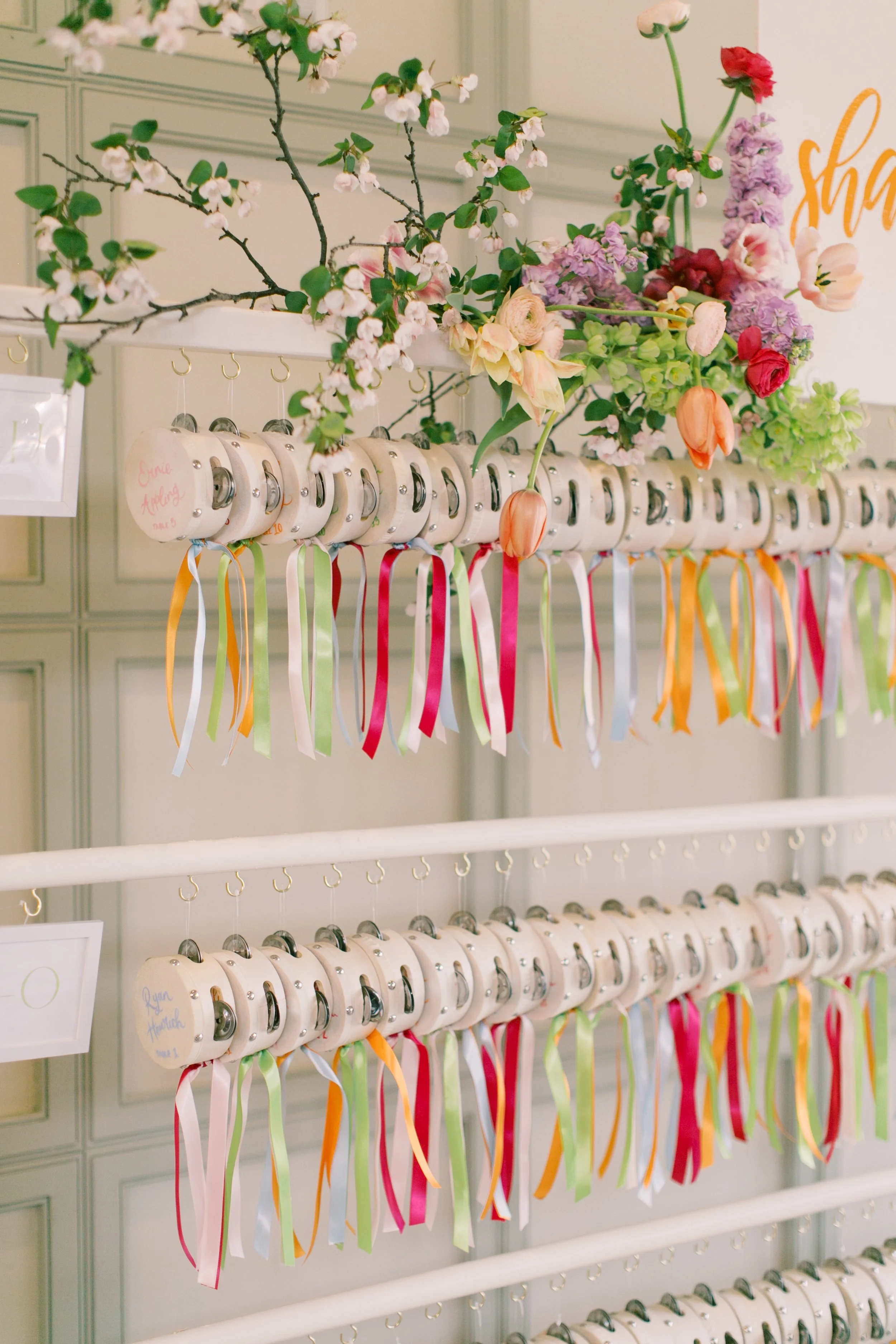 A display of tiny tambourines with colorful ribbons hanging from the handles, decorated with various flowers and branches. Tambourines have handwritten names on them, arranged on white racks against a paneled wall.
