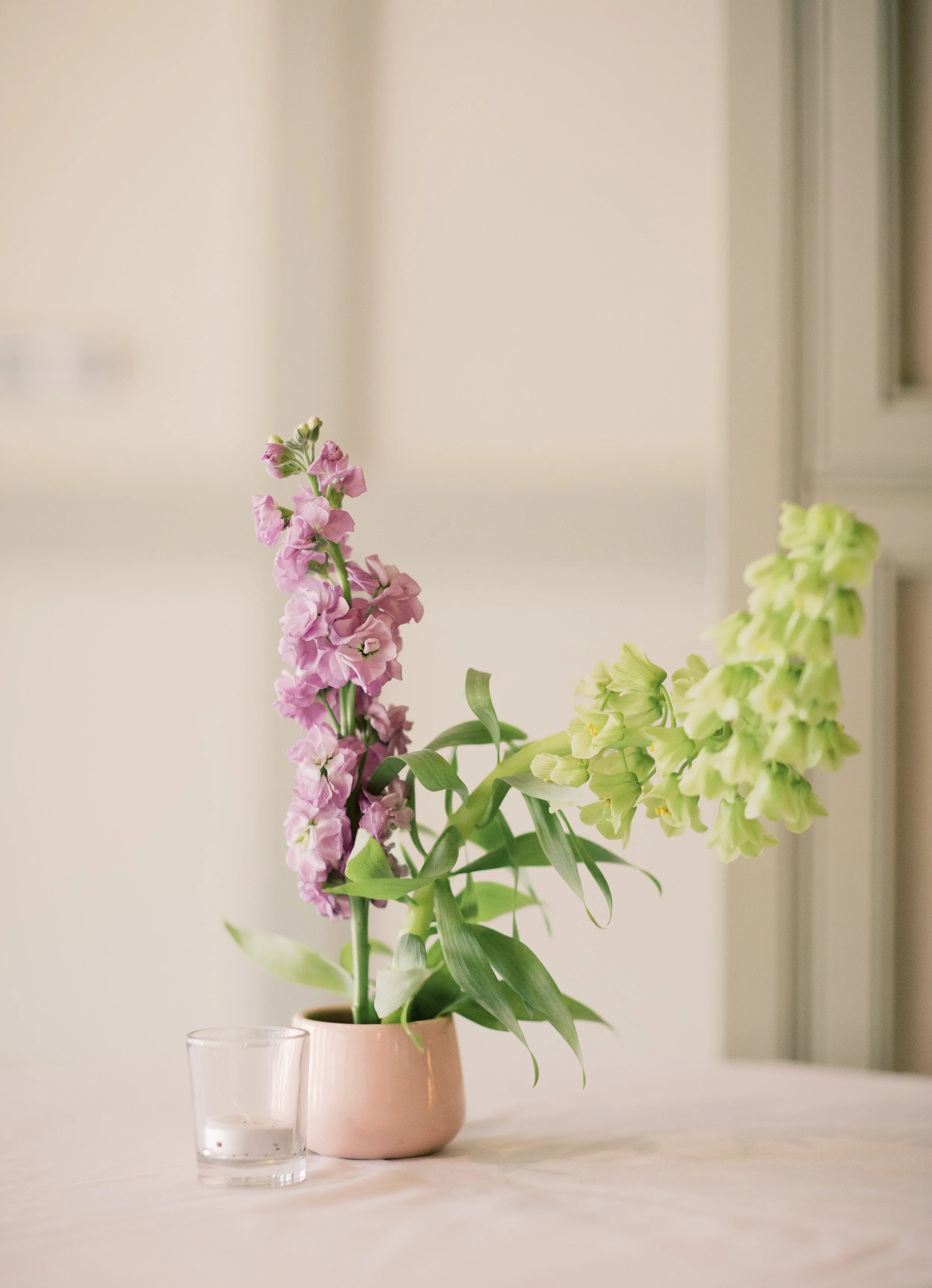 Pink and light yellow flowers in a pink vase on a white table with a small glass of water nearby.