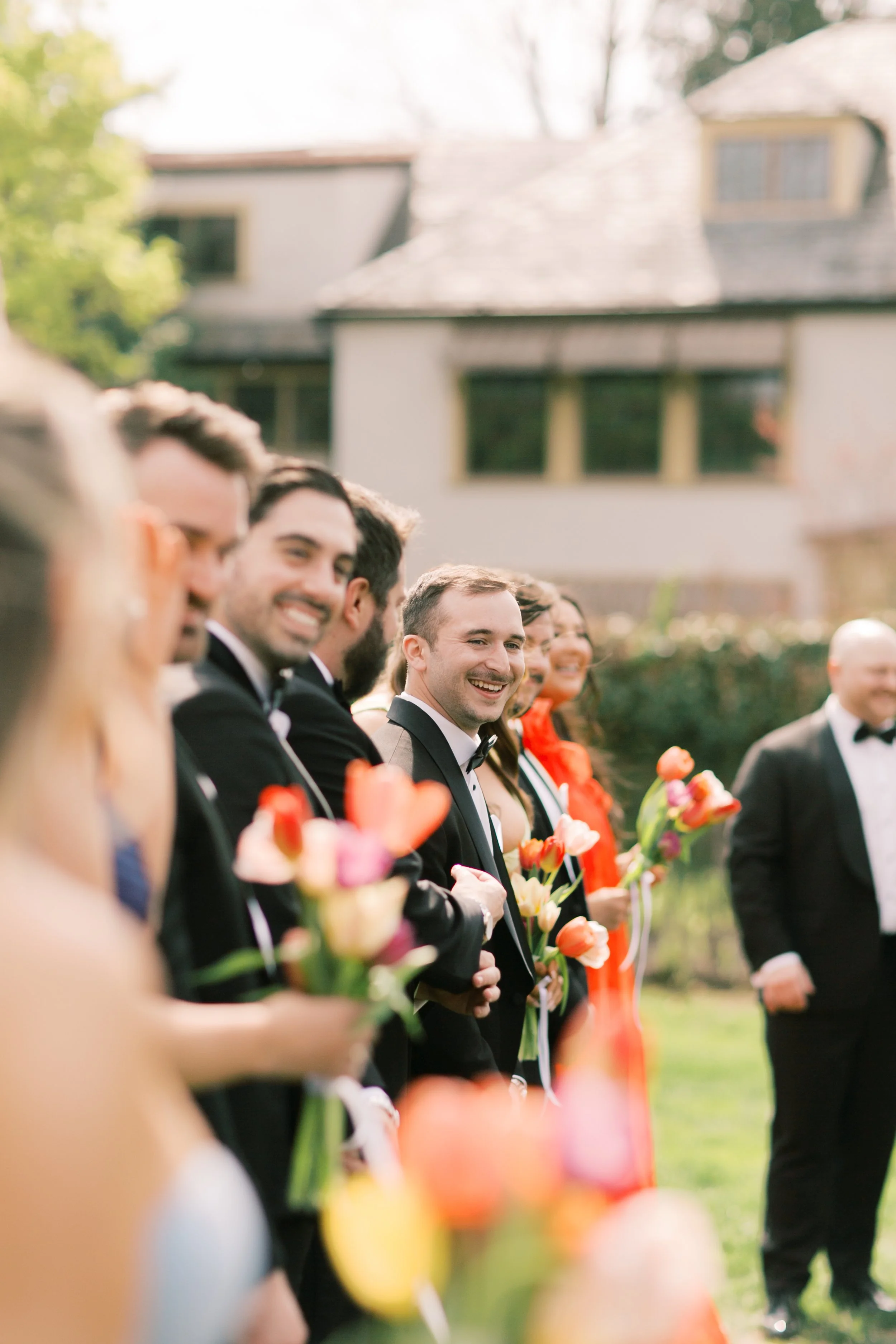 A line of party guests dressed in formal attire holding colorful tulip bouquets, standing outdoors during daytime with a house and trees in the background.