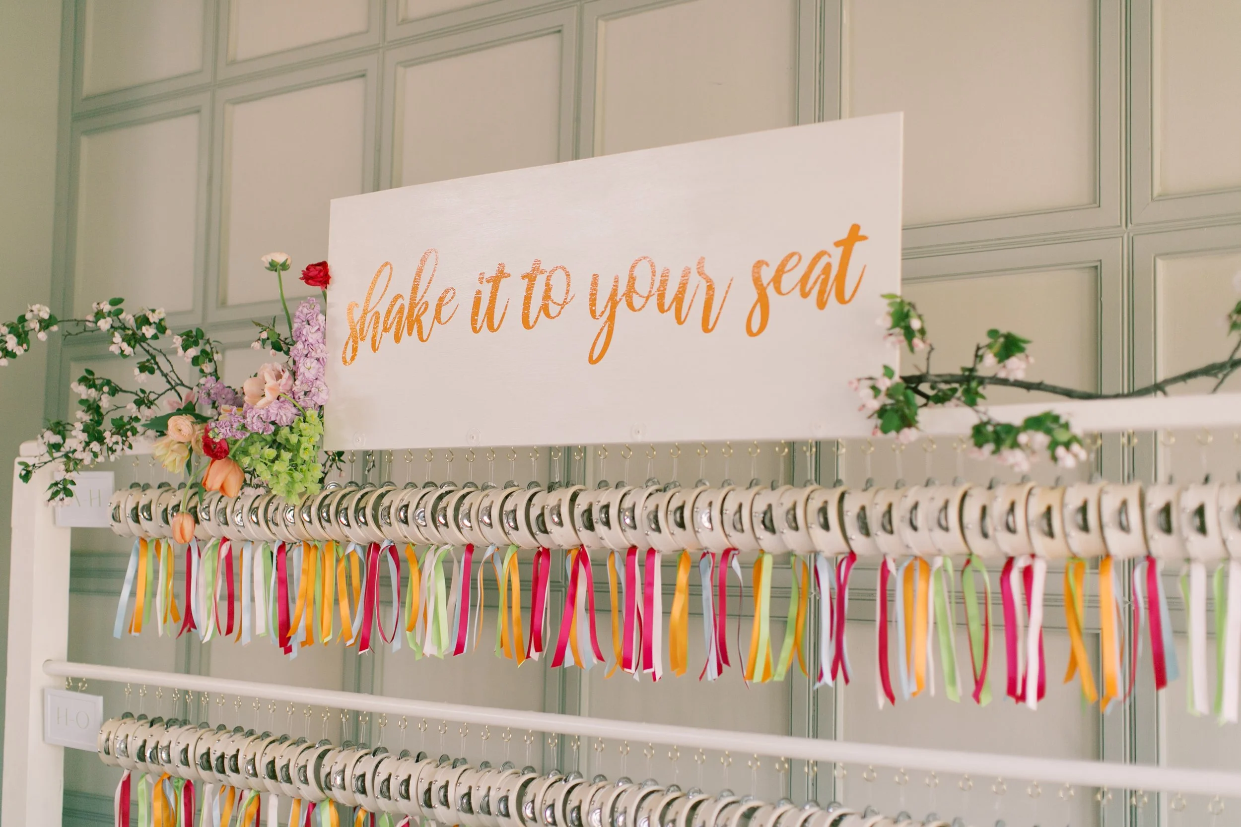 A display of colorful ribbons and rings with a sign that says "shake it to your seat" in orange cursive text, decorated with flowers and branches.