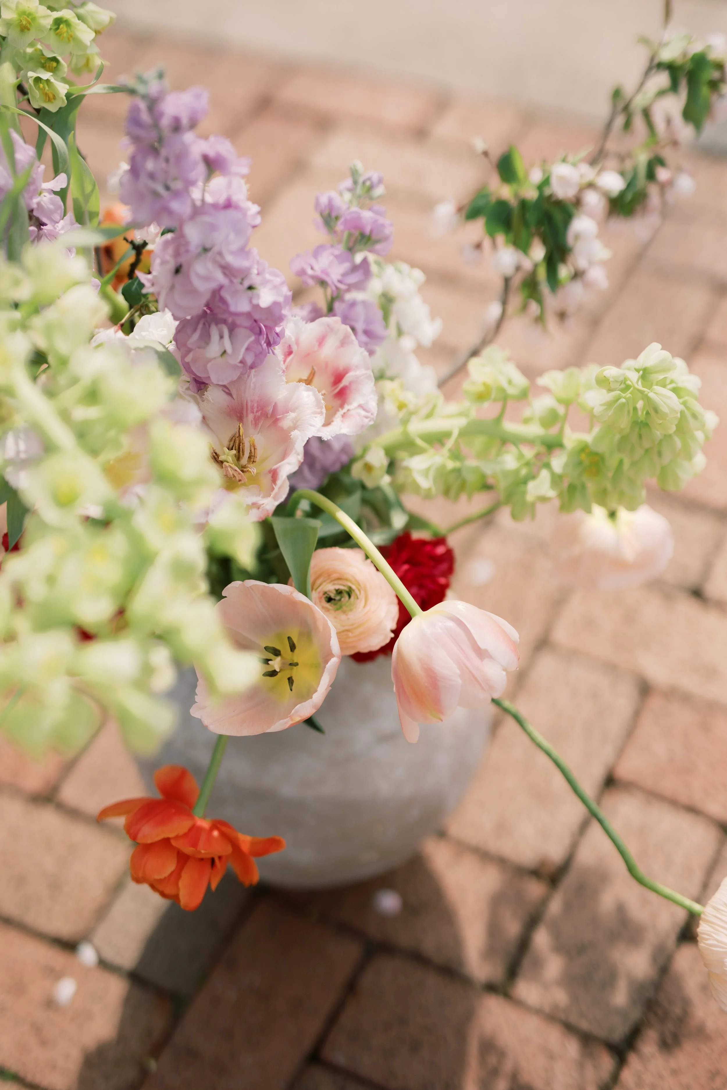 A close-up of a colorful bouquet of mixed tulips and other flowers in a gray pot on brick ground.