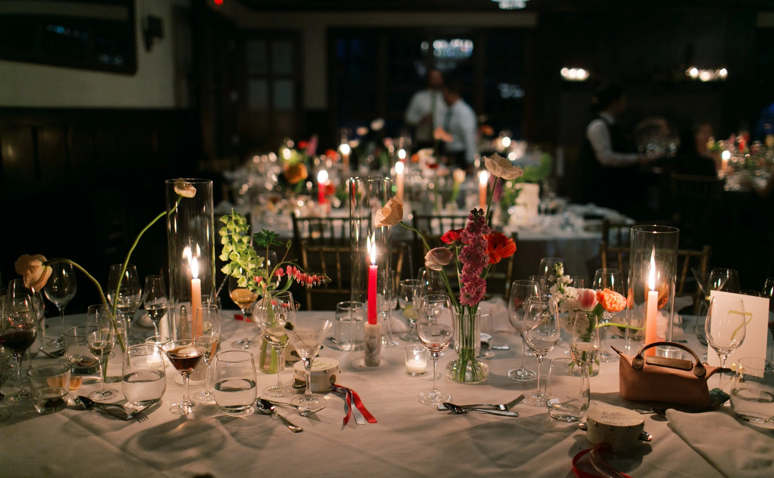 A decorated dinner table set for a celebration, with flowers, candles, glasses, and cutlery, in a dimly lit venue with waitstaff and guests in the background.