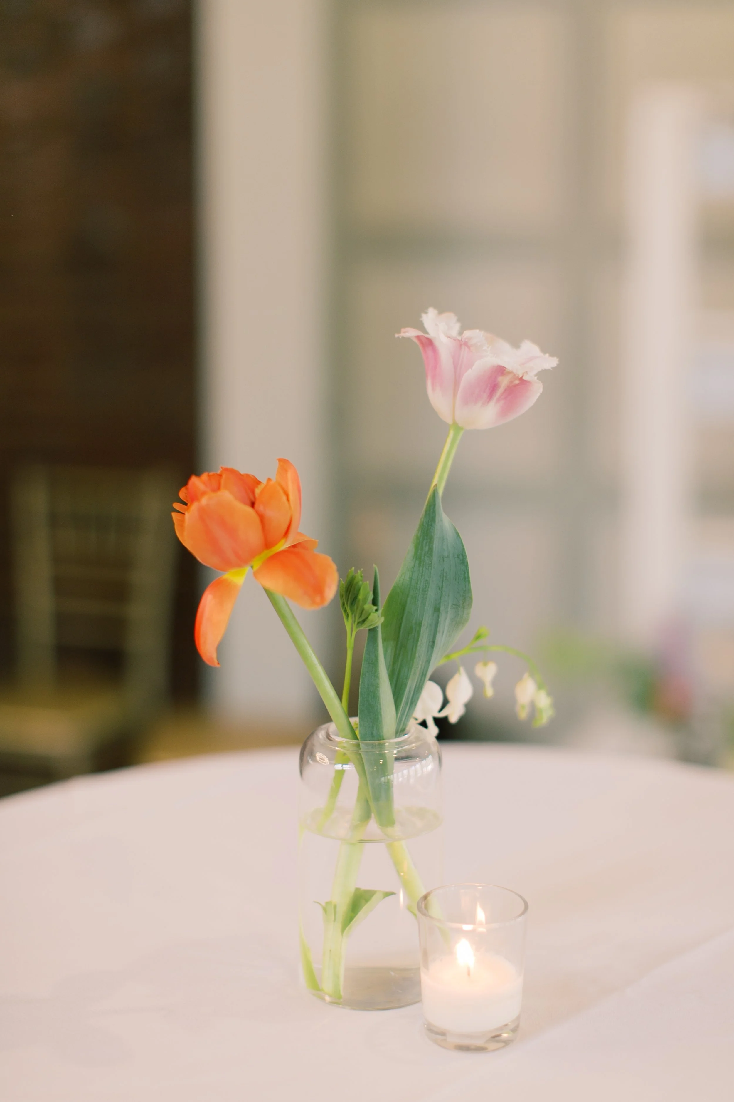 A small glass vase with pink and orange tulips and small white flowers, placed on a white tablecloth, with a lit candle nearby.