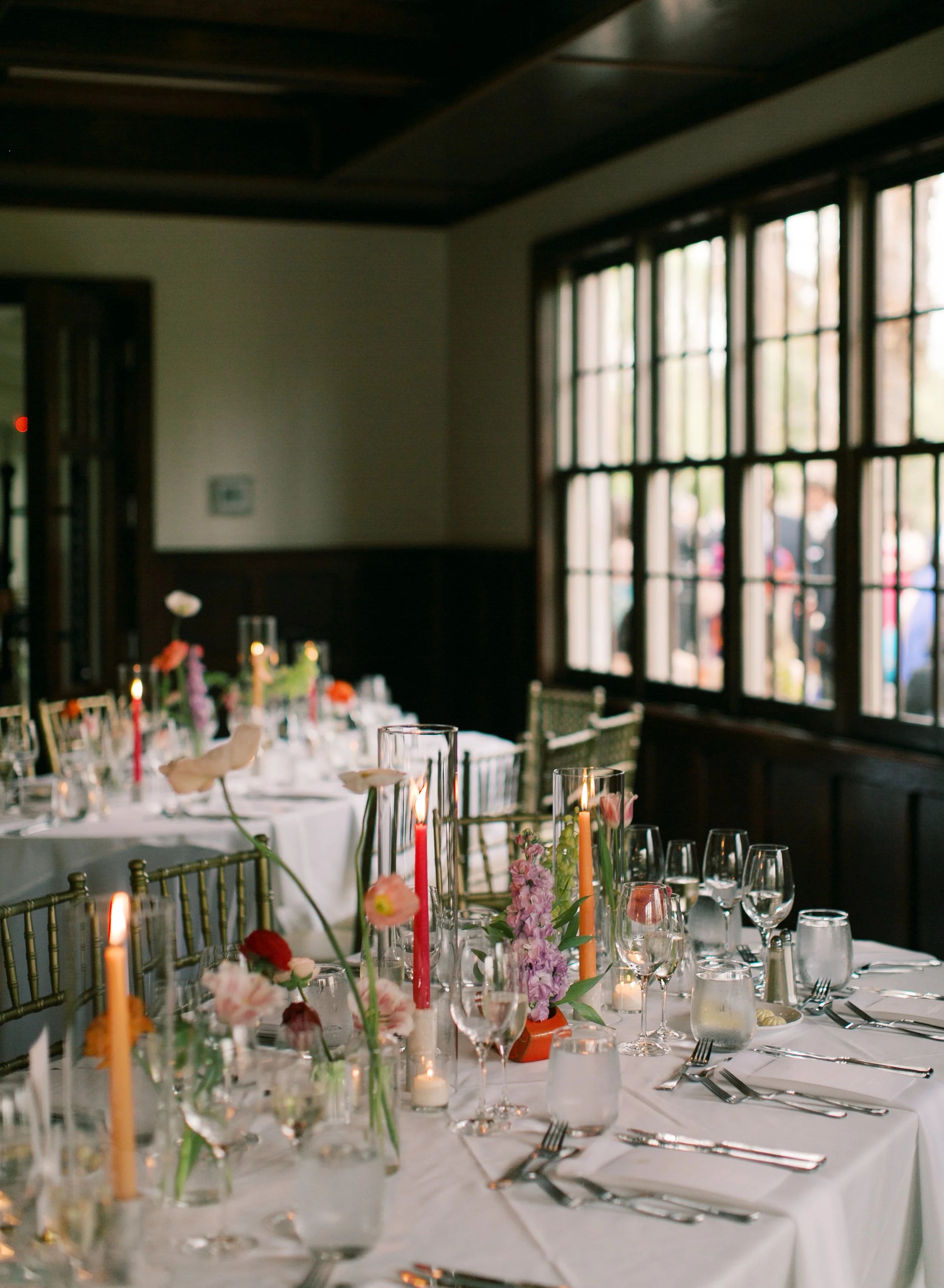 Elegant dining table set for an event with white tablecloth, glassware, cutlery, floral arrangements, and candles inside a room with large windows.