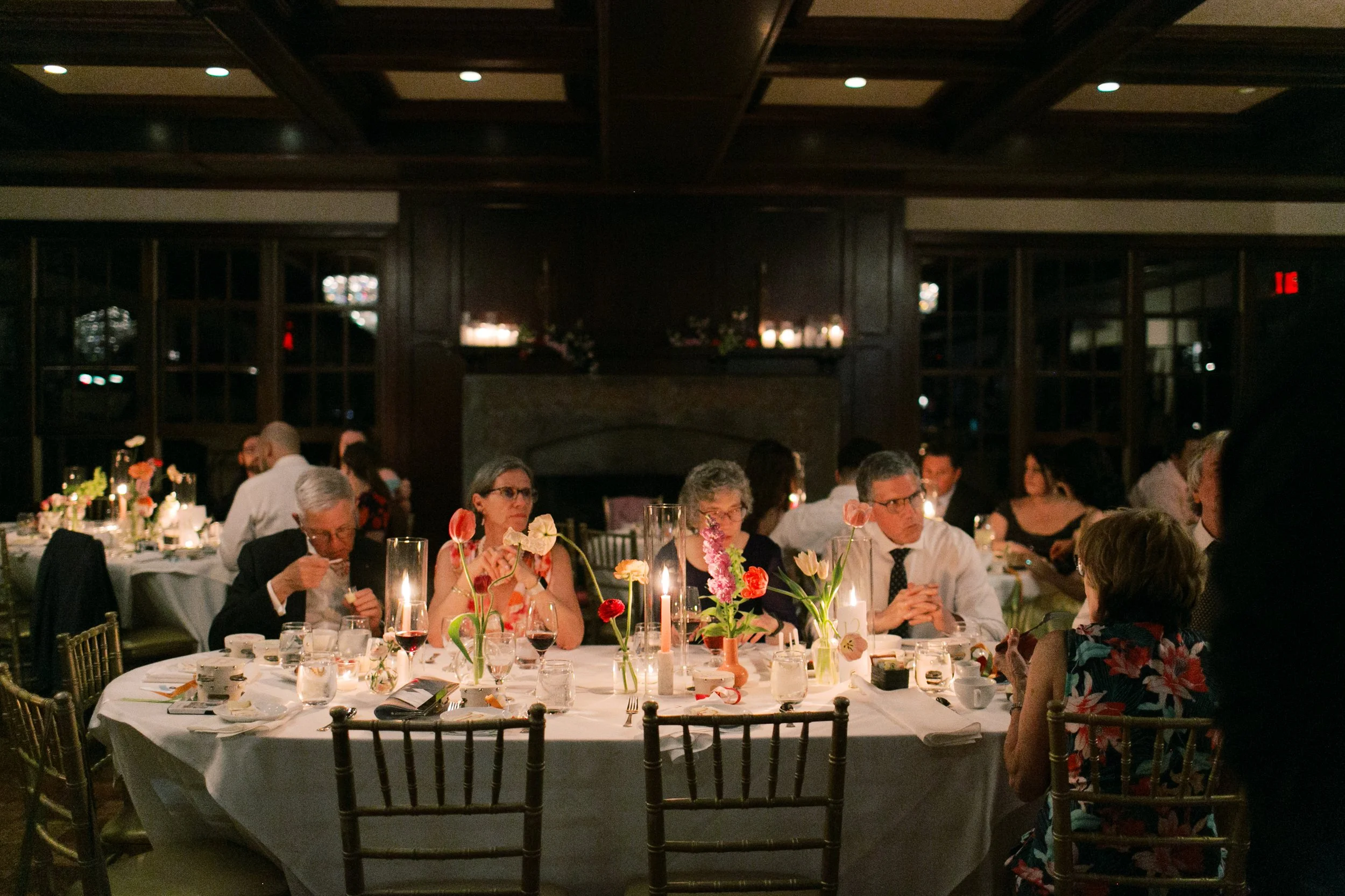 Guests seated at a decorated round table with flowers, candles, and tableware at a formal dinner event in a dimly lit room.