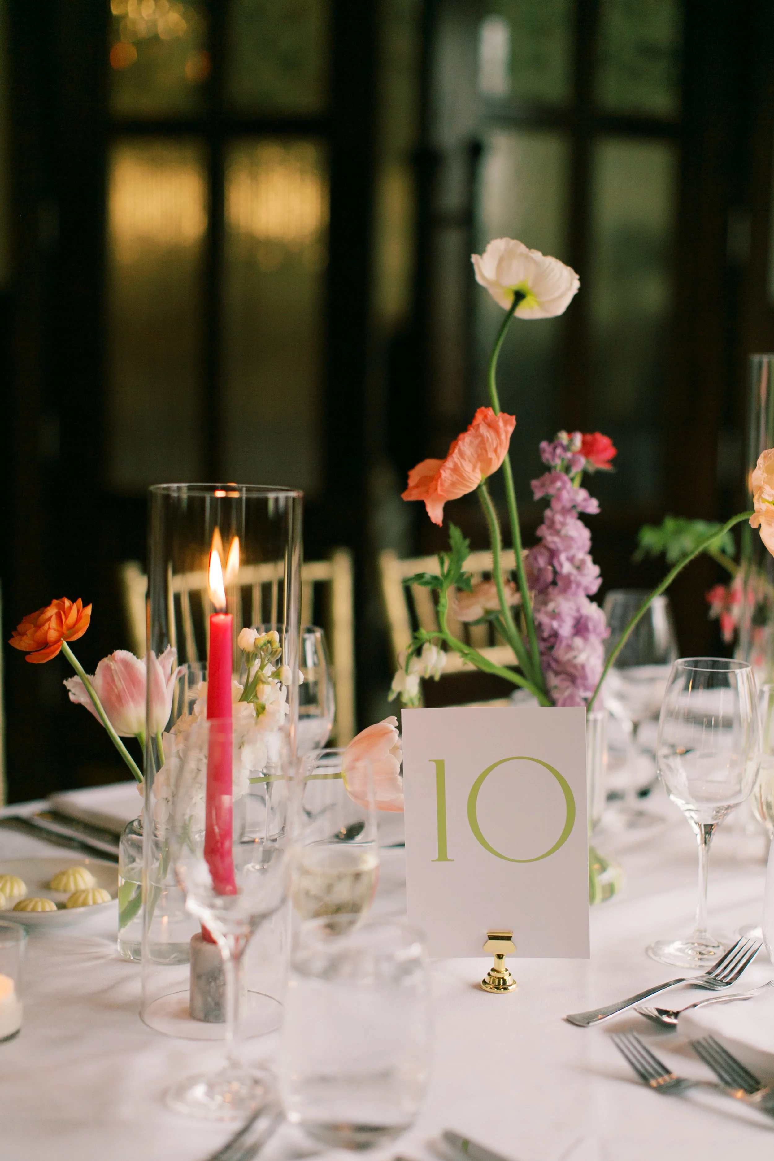 A decorated banquet table with floral arrangements, a lit pink taper candle in a glass holder, and a table number 10 card. The flowers include poppies, roses, and other mixed blooms, arranged in vases, with wine glasses and utensils set around the ta